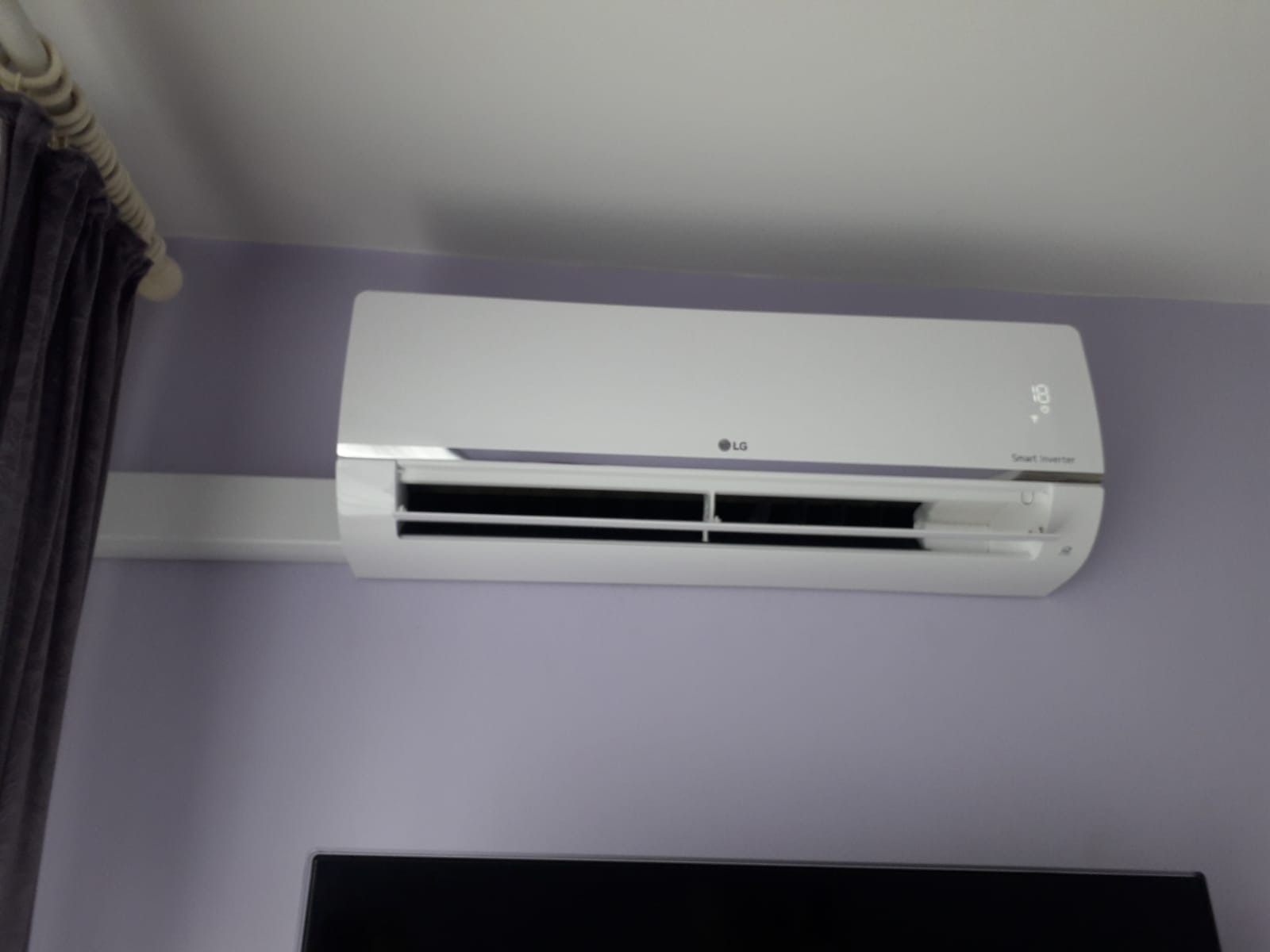 Biała jednostka klimatyzacji LG Smart Inverter zamontowana na fioletowej ścianie, widoczne czarne żaluzje i fragment zasłony po lewej stronie.