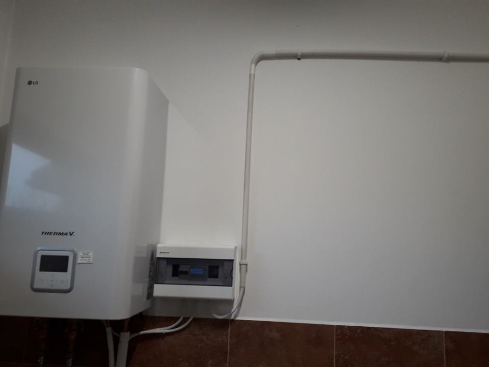 Biała pompa ciepła LG Therma V zamontowana na ścianie z brązowymi płytkami, obok skrzynka elektryczna i rurka instalacyjna.