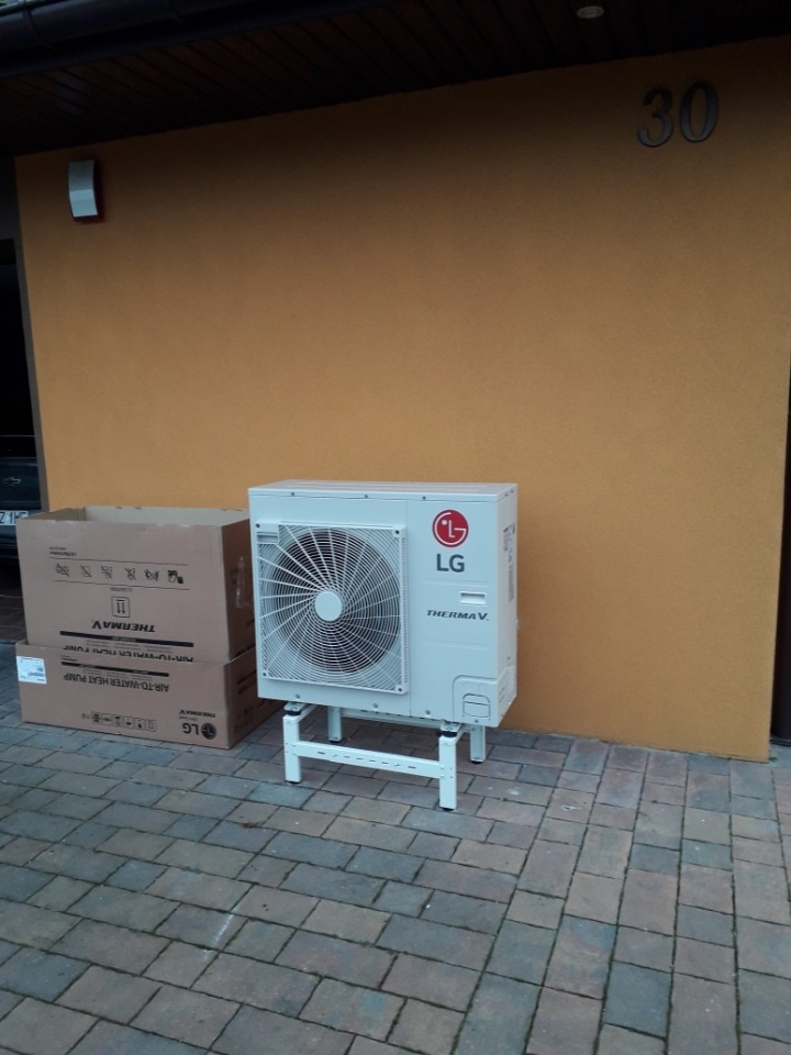 Biała pompa ciepła LG Therma V na białych wspornikach, ustawiona na kostce brukowej przed żółtą ścianą budynku z numerem 30, obok kartonowych pudeł z oznaczeniami producenta.