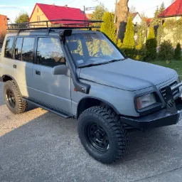 Szary samochód terenowy Suzuki Vitara z orurowaniem, snorkel, bagażnikiem dachowym i oponami terenowymi na tle domów jednorodzinnych.