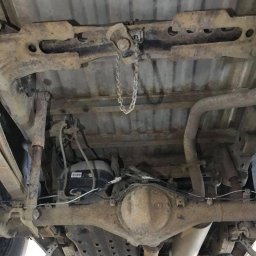 JD Garage - Widok podwozia samochodu z widocznymi elementami zawieszenia, układem wydechowym i zbiornikiem, pokrytymi rdzawym nalotem.