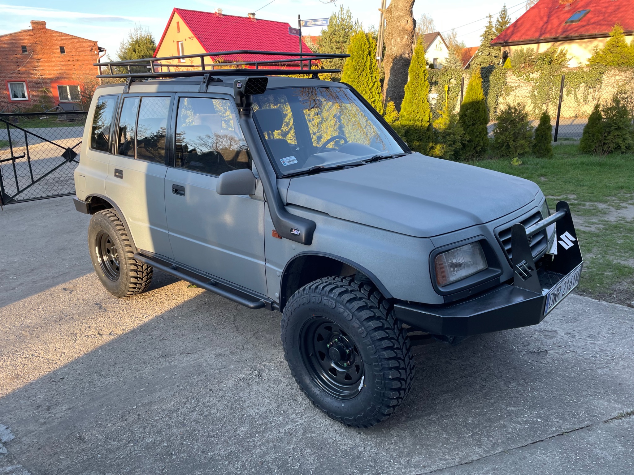 Szary samochód terenowy Suzuki Vitara z orurowaniem, snorkel, bagażnikiem dachowym i oponami terenowymi na tle domów jednorodzinnych.