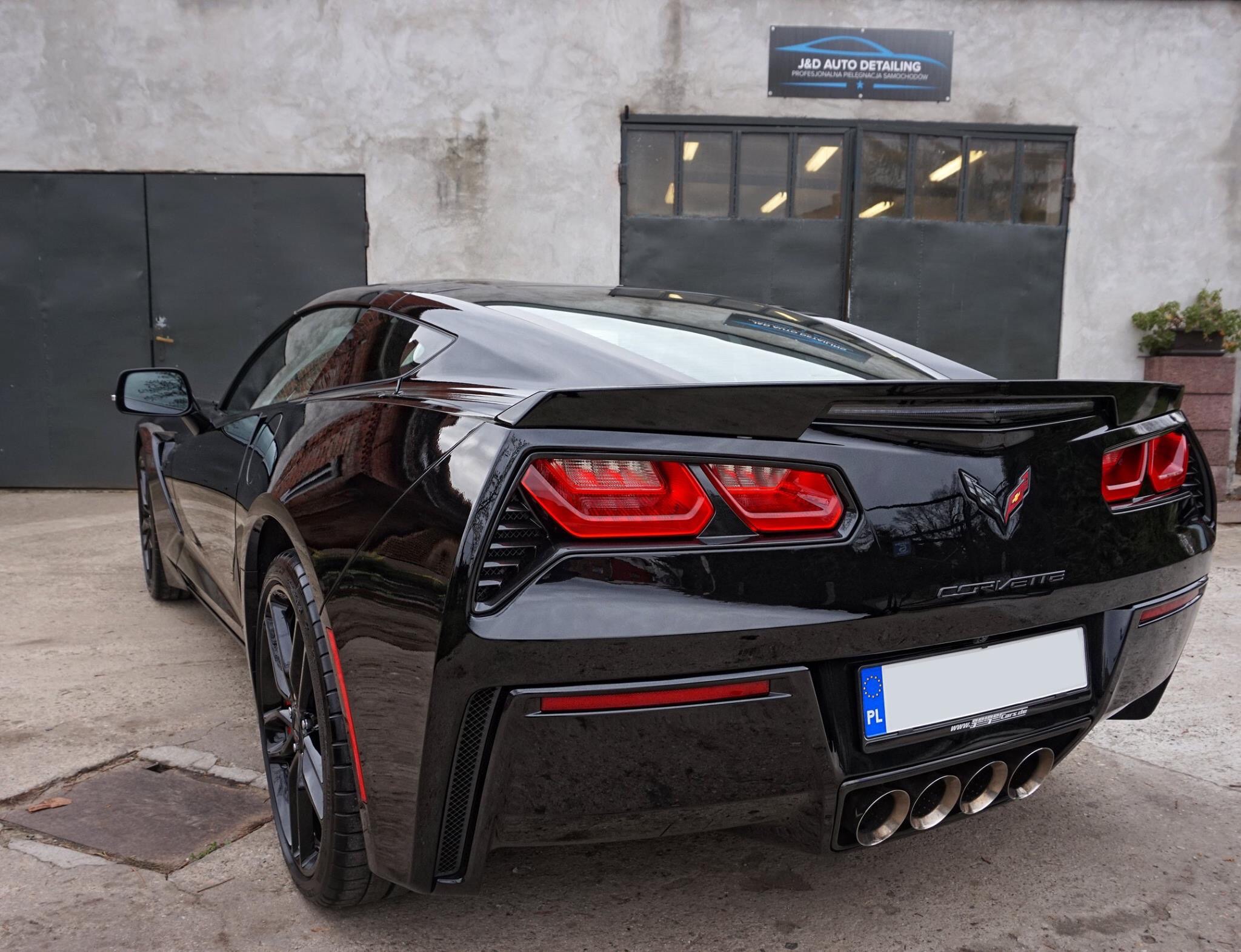 Błyszczący czarny Chevrolet Corvette po detailingu, widok z tyłu na tle szarej ściany budynku z szyldem J&D Auto Detailing.
