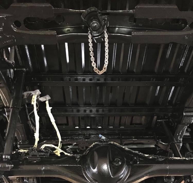 Spód samochodu po zabezpieczeniu antykorozyjnym, widoczne elementy zawieszenia i podwozia pokryte czarną, błyszczącą powłoką, łańcuch podtrzymujący element konstrukcyjny.