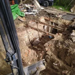Wykop pod fundamenty starego budynku z cegły, widoczny fragment koparki i ATV w tle.