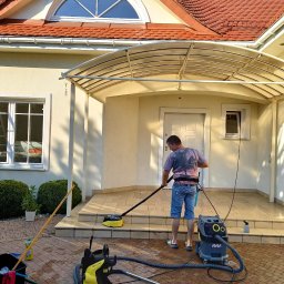 R.M.K Cleaning Service - Mężczyzna w jeansowych spodenkach czyści kostkę brukową przed domem za pomocą myjki ciśnieniowej K5 Full Control i okrągłej szczotki, obok stoi odkurzacz przemysłowy i wiadro z mopem.