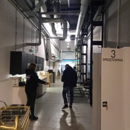 R.M.K Cleaning Service - Wnętrze magazynu lub pomieszczenia gospodarczego z widocznymi instalacjami pod sufitem, osobami i wózkiem transportowym.