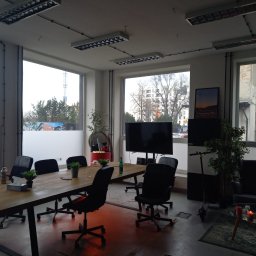 R.M.K Cleaning Service - Jasne, przestronne biuro z dużym drewnianym stołem konferencyjnym, czarnymi krzesłami obrotowymi, roślinami doniczkowymi i oknami z widokiem na miasto. W tle sofa i hulajnoga elektryczna.