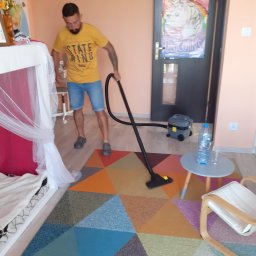 R.M.K Cleaning Service - Mężczyzna w żółtej koszulce odkurza kolorowy dywan w pokoju, widoczny odkurzacz, stolik z butelką wody i plakat z jednorożcami na drzwiach.