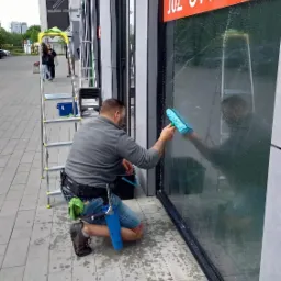 Pracownik na kolanach myje okno budynku z napisem 'JUŻ OTWARTE', używając niebieskiego mopa, obok drabina i wiadro.