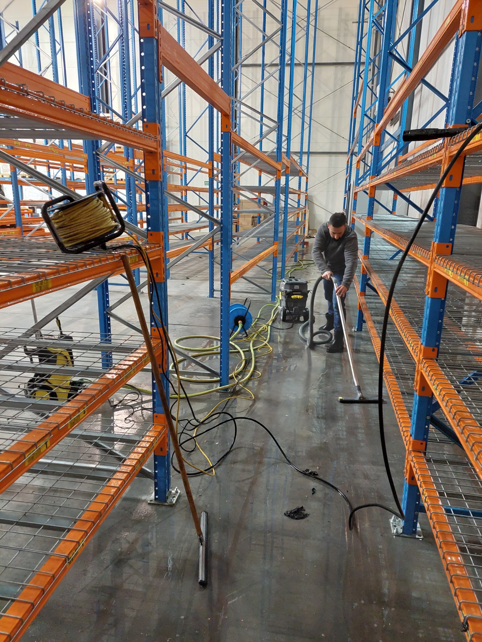 Mężczyzna w szarym swetrze czyści podłogę hali magazynowej przy użyciu odkurzacza przemysłowego, widoczne wysokie regały magazynowe pomalowane na niebiesko i pomarańczowo, żółte kable i węże leżące...