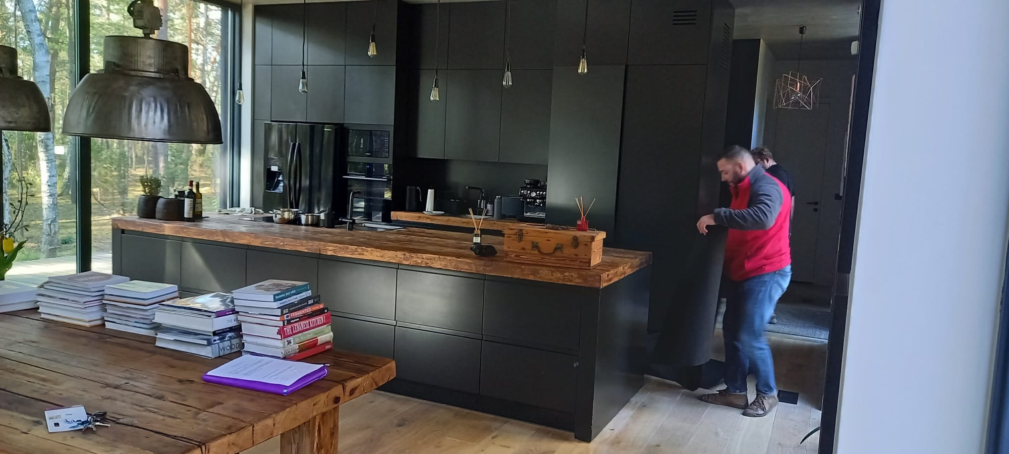 Nowoczesna, czarna kuchnia z drewnianym blatem i industrialnymi lampami, widoczny stos książek na stole i mężczyzna w czerwonej bluzie w tle.