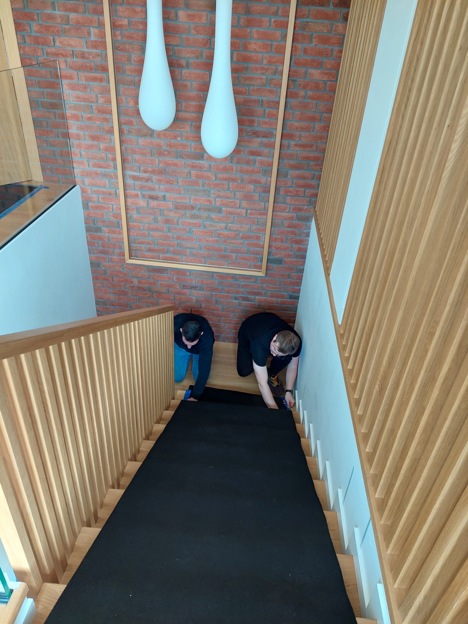 Montaż czarnej wykładziny na drewnianych schodach w nowoczesnym wnętrzu z ceglaną ścianą i designerskim oświetleniem. Dwóch mężczyzn precyzyjnie docina materiał przy listwach.