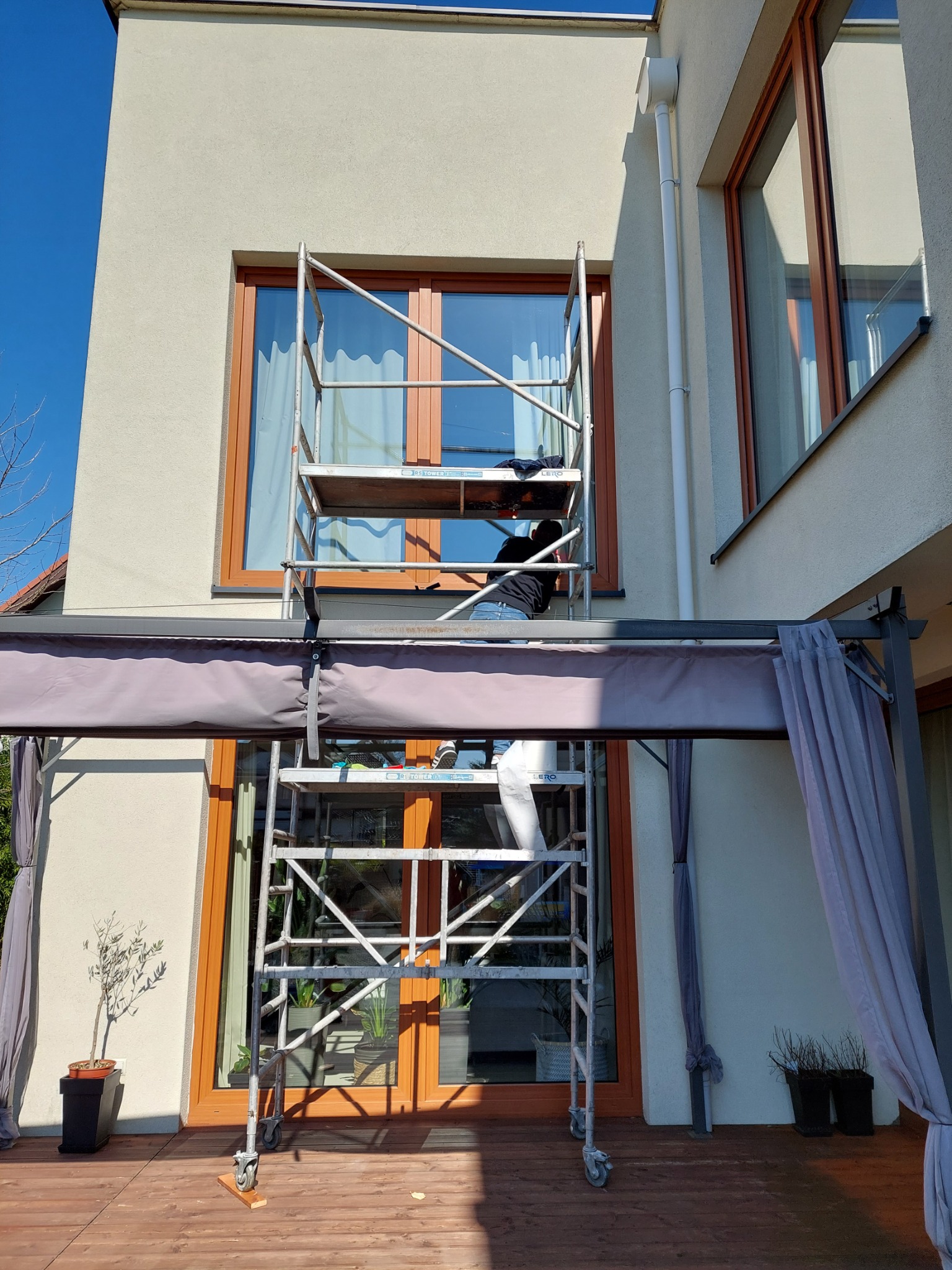 Pracownik na rusztowaniu myje okno na wysokości, widoczny fragment elewacji domu i drewniany taras z pergolą.