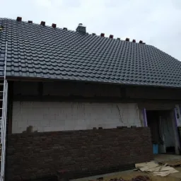 Fragment dachu z czarnej dachówki karpiówki, częściowo pokryty, z widocznym kominem, oraz fragment ściany budynku w trakcie murowania z cegły klinkierowej, z widoczną drabiną przy dachu.