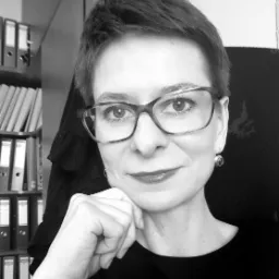 Portret kobiety w okularach, z ręką pod brodą, na tle półki z segregatorami w czarno-białej tonacji.