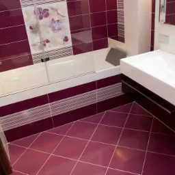 Wnętrze łazienki z bordowymi płytkami podłogowymi ułożonymi w karo, bordowe płytki ścienne z motywem kwiatowym nad wanną z parawanem oraz biała, prostokątna umywalka.