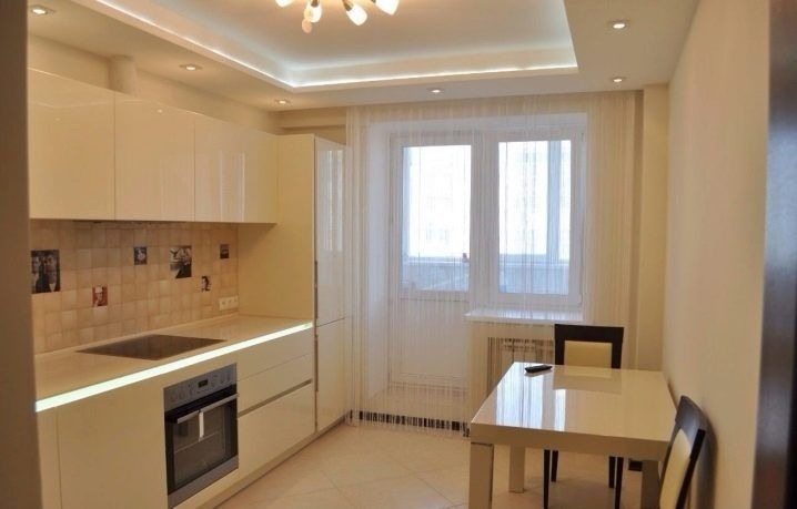 Jasna kuchnia z białymi, lakierowanymi szafkami, ceramiczną mozaiką z czarno-białymi zdjęciami nad blatem, oraz białym stołem z ciemnymi krzesłami w pobliżu okna z firanką sznurkową.