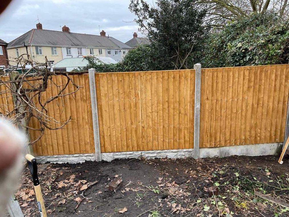 Nowo postawione drewniane ogrodzenie panelowe z betonowymi słupkami i podmurówką na nierównym terenie, z widoczną łopatą i pozostałościami po pracach ziemnych.