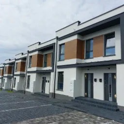 Szereg nowoczesnych domów z białą elewacją, drewnianymi panelami i szarymi elementami wykończeniowymi, widoczne skrzynki gazowe i drabina na elewacji jednego z domów. Osiedle Szeregówek przy ul.