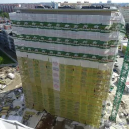 Wysoki budynek w trakcie ocieplania, osłonięty siatką ochronną z logo Greinplast, widok z góry na plac budowy z dźwigiem i materiałami izolacyjnymi.