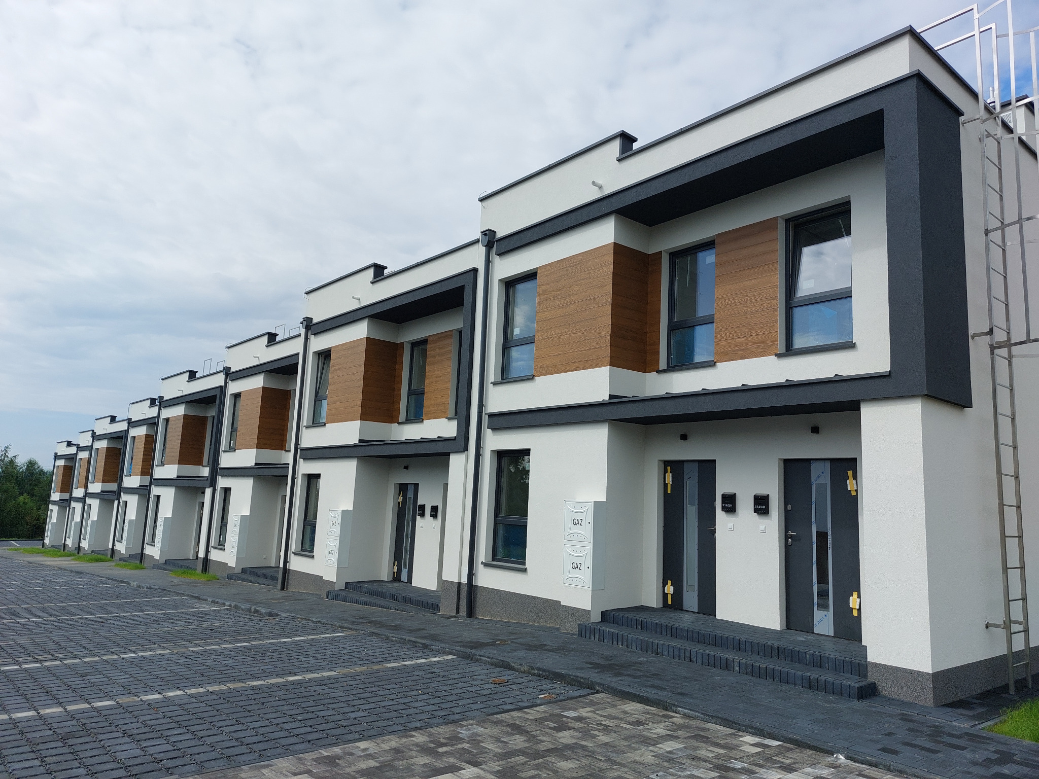 Szereg nowoczesnych domów z białą elewacją, drewnianymi panelami i szarymi elementami wykończeniowymi, widoczne skrzynki gazowe i drabina na elewacji jednego z domów. Osiedle Szeregówek przy ul.