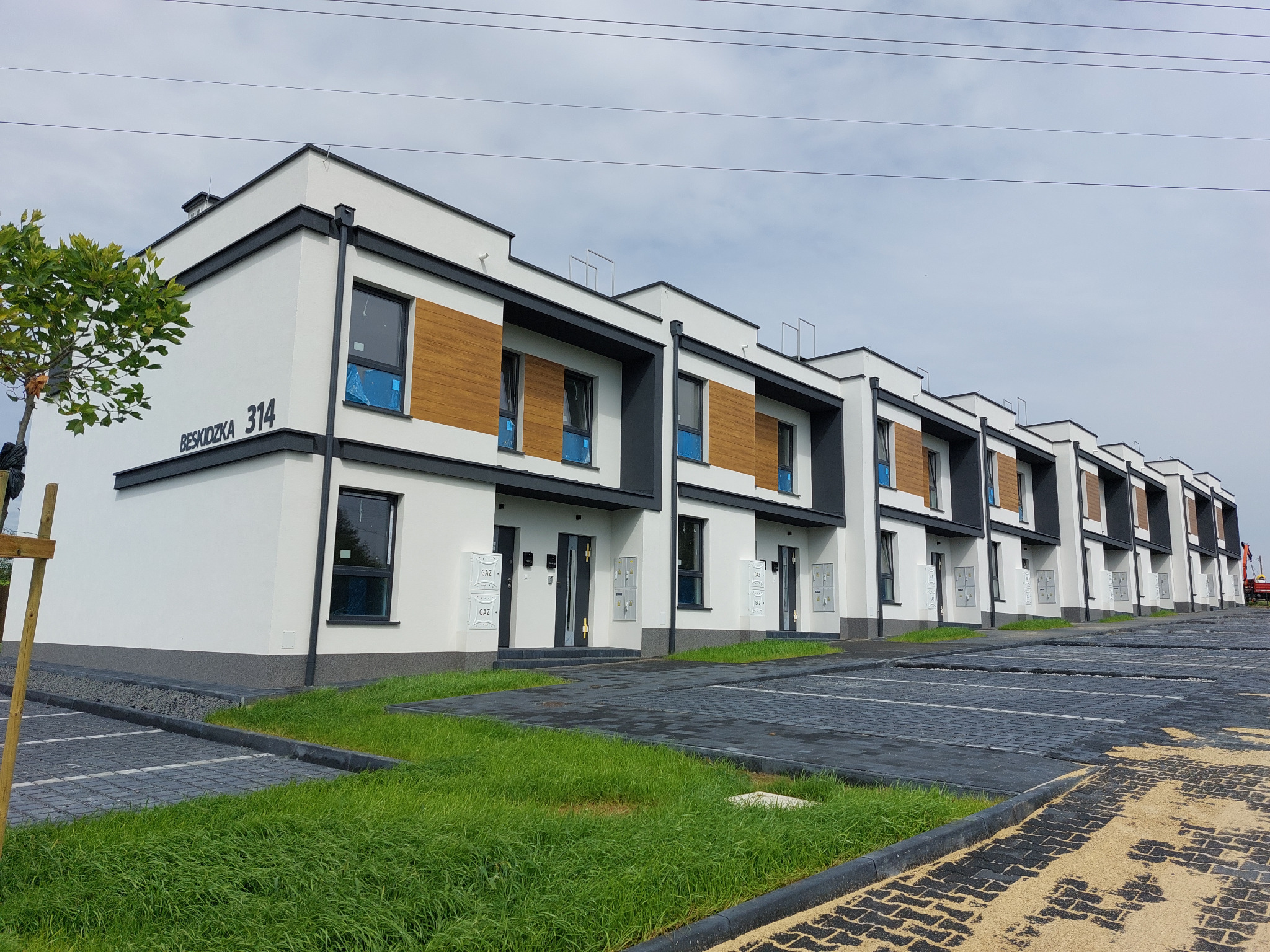 Nowoczesne szeregówki z białymi elewacjami, drewnianymi panelami i szarymi akcentami, widok z ulicy Beskidzkiej, świeżo posadzona zieleń i wybrukowany parking.