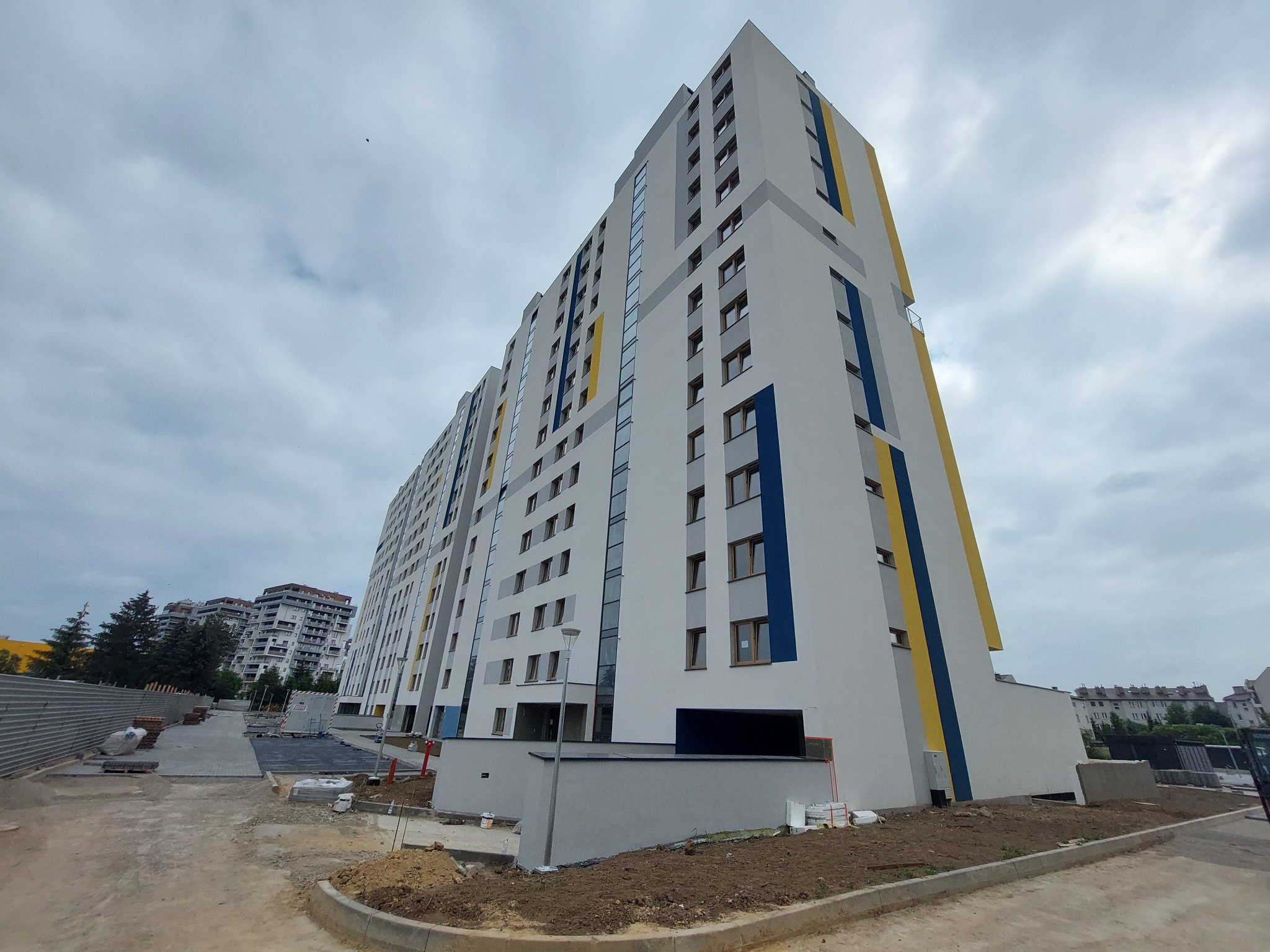 Nowoczesny blok mieszkalny z szarą elewacją, akcentami w kolorze niebieskim i żółtym, widok z dołu na Osiedlu Nova Graniczna, teren w trakcie zagospodarowania.