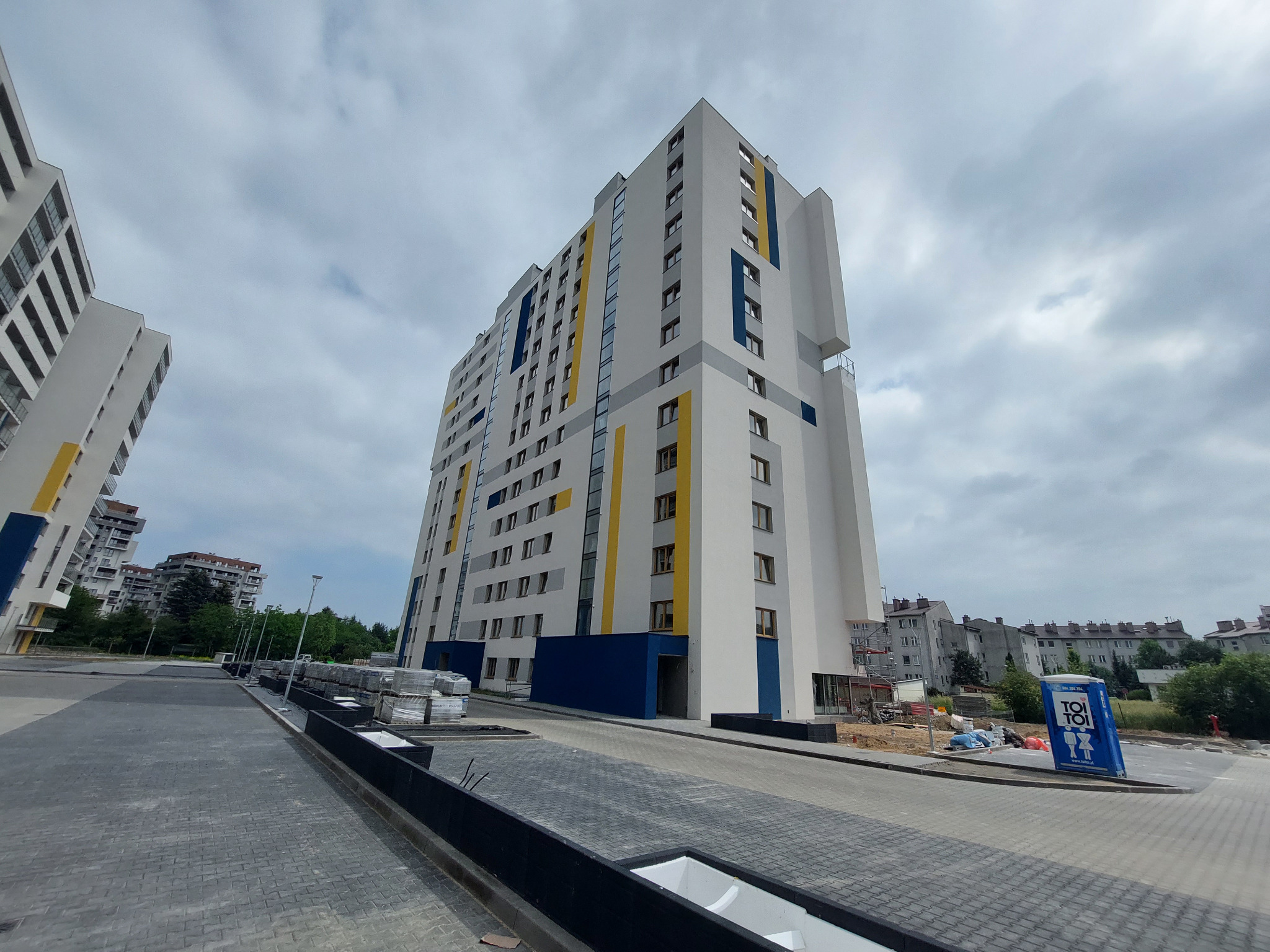 Wielopiętrowy budynek mieszkalny z białą elewacją, urozmaiconą pionowymi pasami w kolorach żółtym i niebieskim, widoczny z poziomu chodnika z kostki brukowej, z toaletą przenośną TOI TOI w tle...
