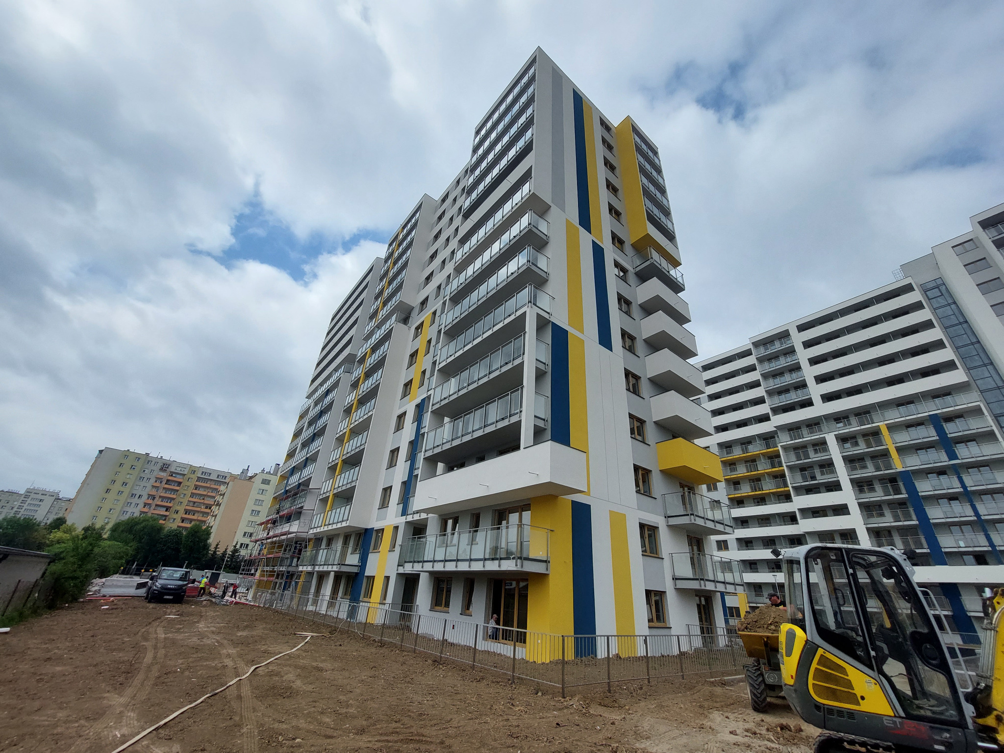 Nowoczesny apartamentowiec z balkonami, pomalowany w pasy żółte i niebieskie, widoczny z dołu, z minikoparką na pierwszym planie i elementami rusztowania po lewej stronie.