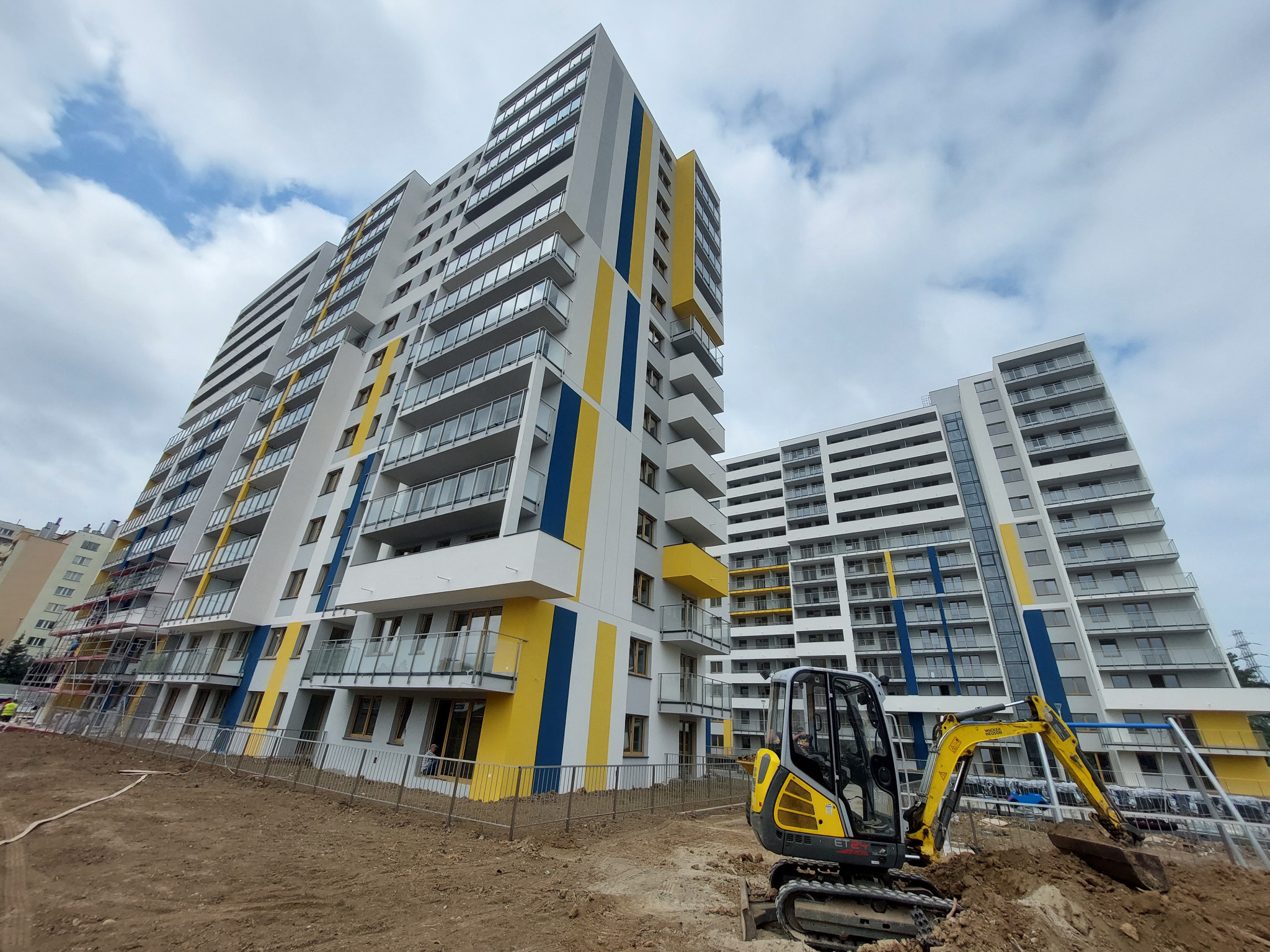Nowoczesny budynek mieszkalny z kolorową elewacją (żółte i niebieskie pasy) w trakcie budowy, widoczny mały żółty koparka na pierwszym planie, ziemia rozkopana, pochmurne niebo.
