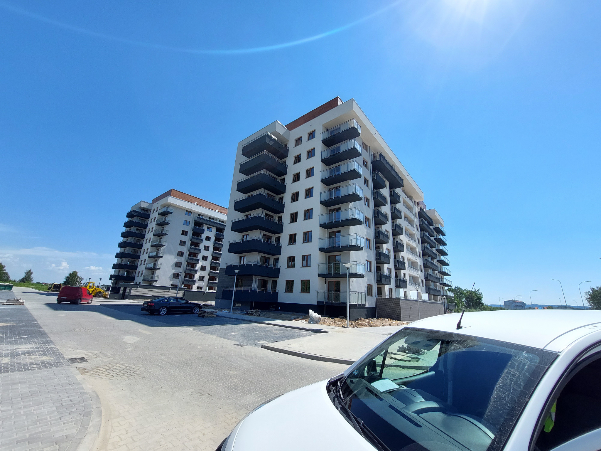 Dwa nowoczesne, wielopiętrowe budynki mieszkalne z białymi elewacjami i ciemnymi balkonami, widoczne spod niskiego kąta z perspektywy parkingu, z fragmentem białego samochodu dostawczego...