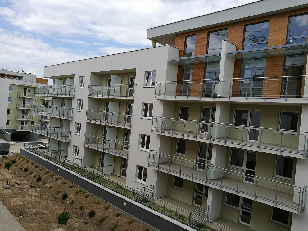 Nowoczesny budynek wielorodzinny z jasną elewacją, szklanymi balustradami balkonów i elementami drewnianymi, widok z góry na zagospodarowywany teren.