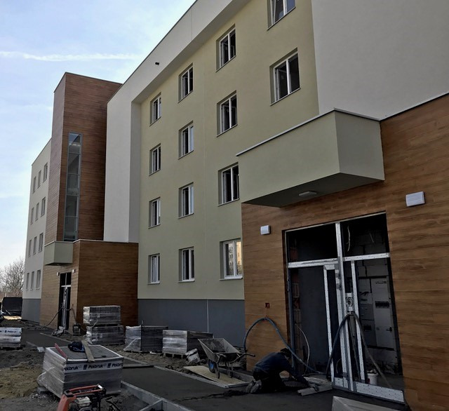 Budynek wielorodzinny w trakcie wykańczania elewacji, widoczne płyty elewacyjne imitujące drewno, ułożone palety z materiałami budowlanymi oraz pracownik układający chodnik z kostki brukowej przed...