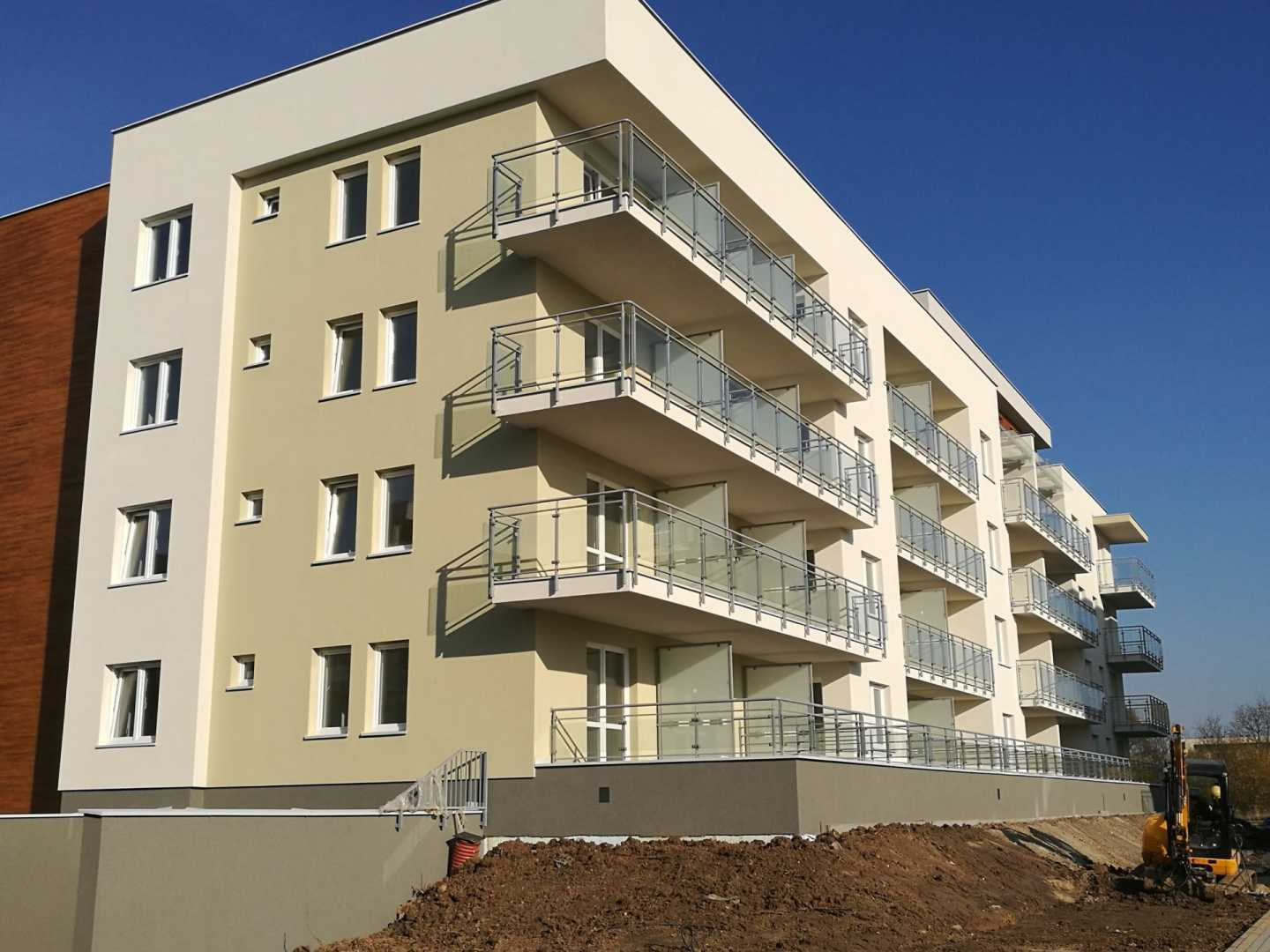 Nowoczesny budynek wielorodzinny z balkonami wykończonymi szkłem i metalem, świeżo po pracach elewacyjnych, z widocznymi śladami ziemi po pracach ziemnych i małą koparką w tle.