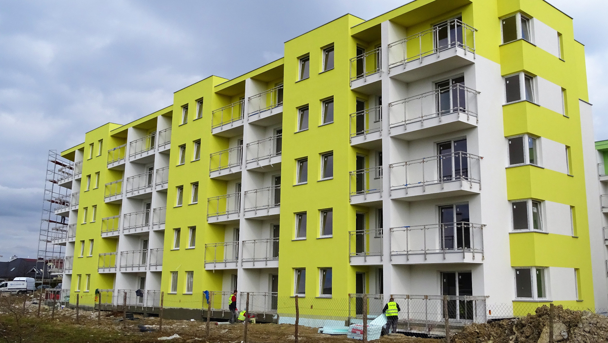 Nowo wybudowany, wielopiętrowy budynek mieszkalny z jasnożółtą elewacją i białymi balkonami z szklanymi balustradami, częściowo otoczony siatką ogrodzeniową, z widocznymi pracownikami...