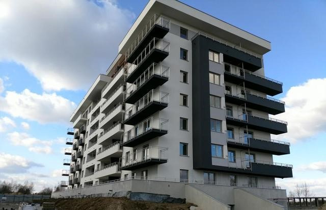 Nowoczesny budynek wielorodzinny z białymi ścianami i ciemnymi balkonami pod błękitnym niebem.