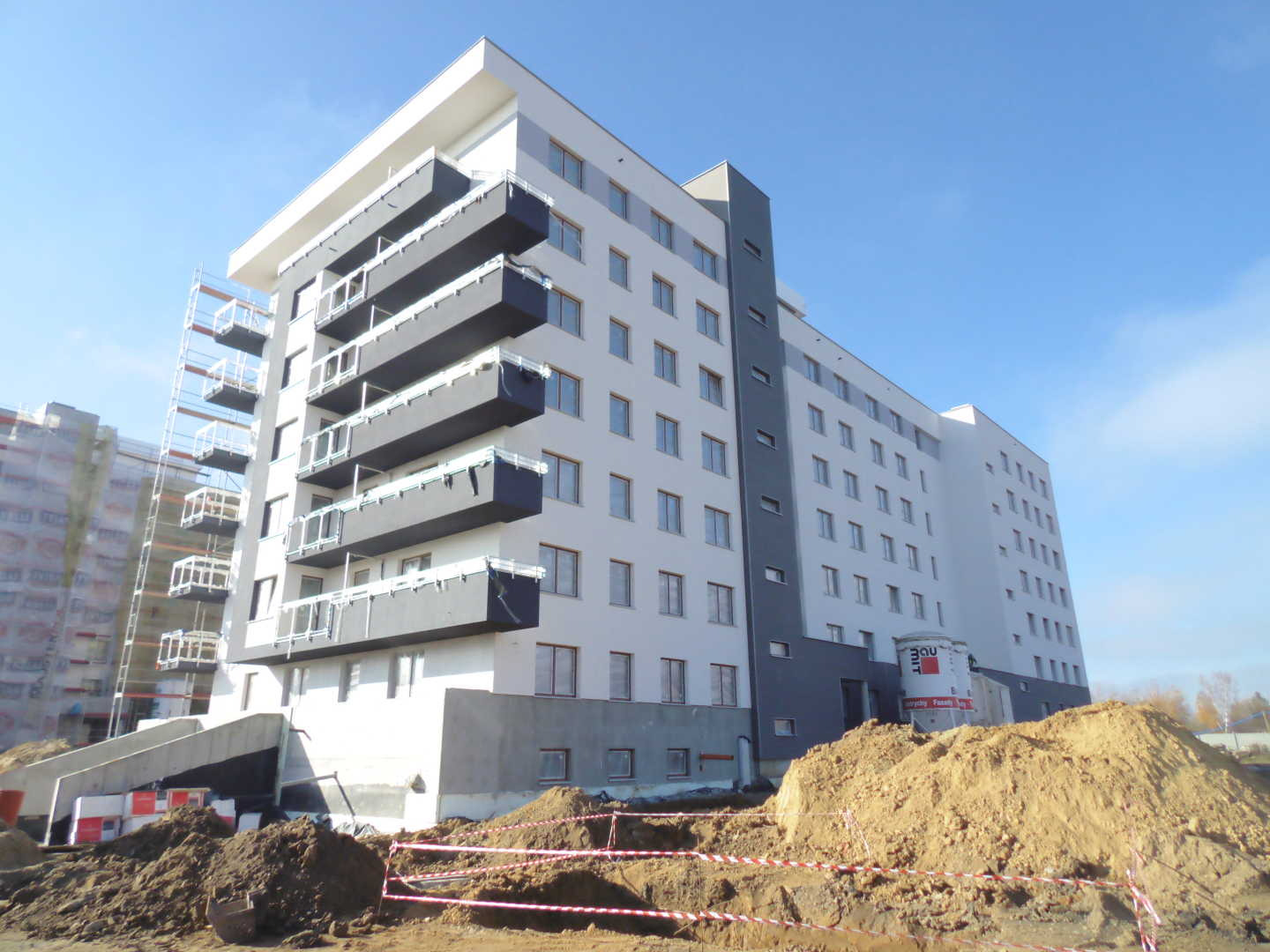 Nowoczesny budynek mieszkalny w trakcie wykończenia elewacji, z widocznymi rusztowaniami, stertą ziemi i materiałami budowlanymi na pierwszym planie.