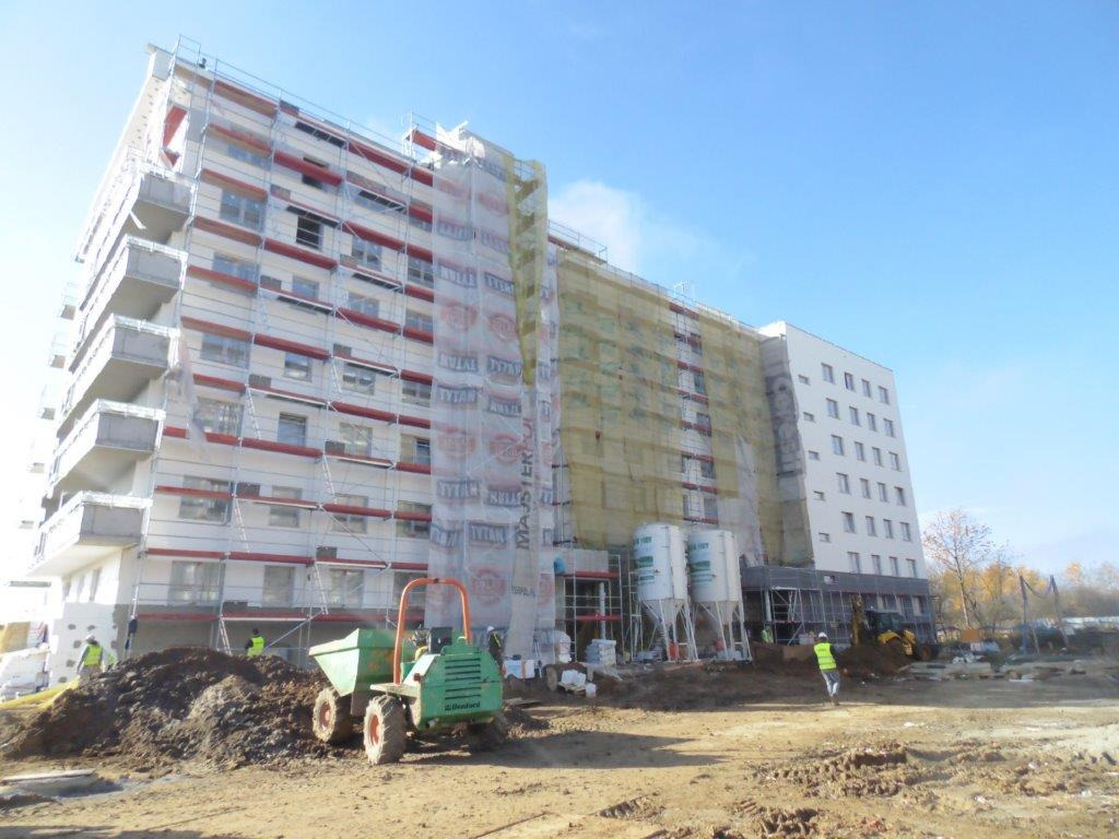 Wielopiętrowy budynek w trakcie prac elewacyjnych, z widocznymi rusztowaniami, siatkami ochronnymi i sprzętem budowlanym na placu budowy; zielona wywrotka na pierwszym planie.