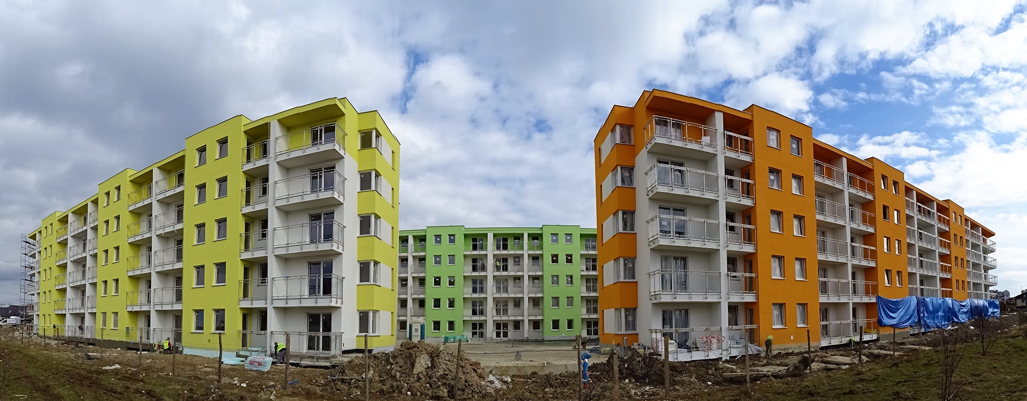 Panorama nowo wybudowanych bloków mieszkalnych o elewacjach w kolorach żółtym, pomarańczowym i zielonym, z widocznymi balkonami z barierkami ze stali nierdzewnej, na pierwszym planie teren budowy...