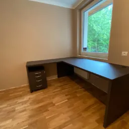 Nowoczesne biurko narożne z ciemnego drewna, z szafką na kółkach, umieszczone przy oknie z widokiem na zieleń, na podłodze parkiet.
