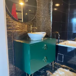 Nowoczesna łazienka z zieloną szafką pod umywalką, złotymi nóżkami i okrągłymi uchwytami, czarne płytki z fakturą na ścianie, okrągłe lustro z odbiciem grzejnika i lamp, marmurowa podłoga.