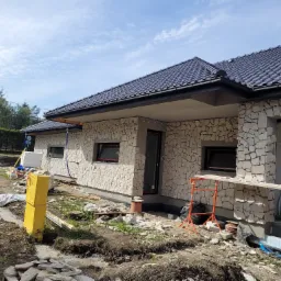 Dom jednorodzinny w trakcie wykończenia elewacji kamieniem naturalnym, widoczne rusztowanie i materiały budowlane, przygotowany teren wokół budynku.