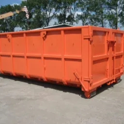 Pomarańczowy kontener na gruz lub odpady budowlane, widoczny na placu budowy z dźwigiem w tle, świeżo po malowaniu.