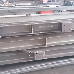 Stalowe belki konstrukcyjne ułożone jedna na drugiej, z widocznymi oznaczeniami i przyspawanymi elementami mocującymi z otworami na śruby.