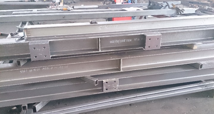 Stalowe belki konstrukcyjne ułożone jedna na drugiej, z widocznymi oznaczeniami i przyspawanymi elementami mocującymi z otworami na śruby.