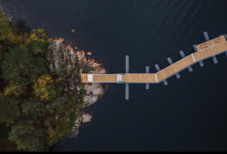 Widok z lotu ptaka na drewniane molo z szarymi podporami, prowadzące od skalistego brzegu porośniętego drzewami w ciemną wodę.