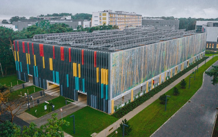 Wielopoziomowy parking z kolorowymi pionowymi panelami (czerwony, żółty, turkusowy) i abstrakcyjnym malowidłem imitującym las, otoczony zielenią i innymi budynkami.