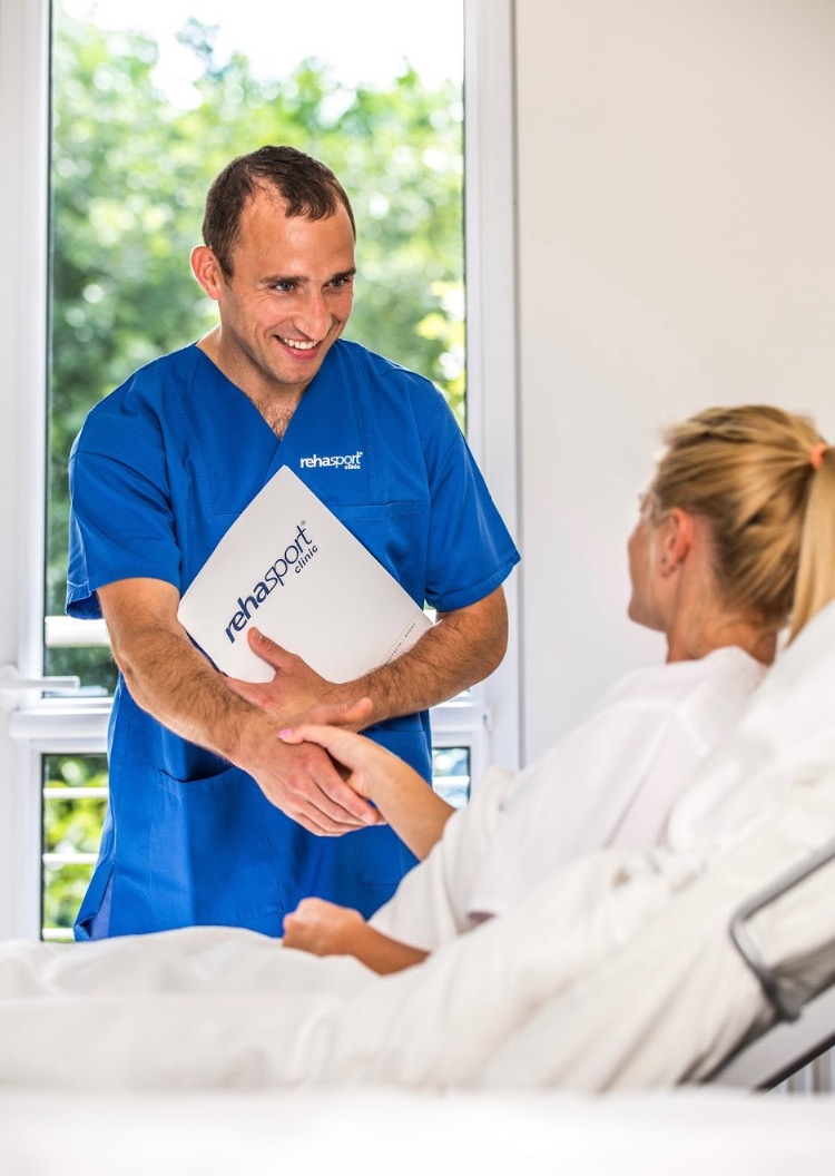 Lekarz w niebieskim uniformie uśmiecha się i podaje rękę pacjentce leżącej w łóżku szpitalnym, trzymając w drugiej ręce folder z logo 'rehasport clinic', za nimi okno z widokiem na zieleń.