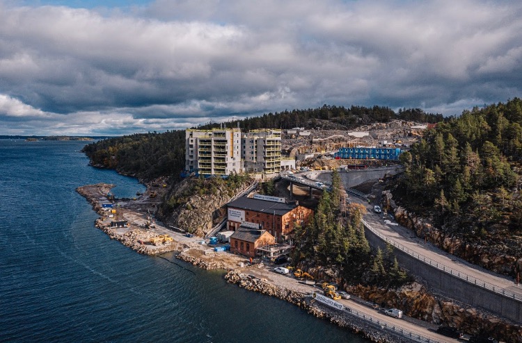 Widok z lotu ptaka na plac budowy nad brzegiem morza, z budynkami w stanie surowym, ciężkim sprzętem i budynkiem przemysłowym z cegły u podnóża wzgórza.